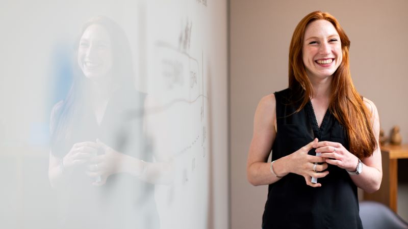 A white female teacher stands at the front of a classroom in front of a whiteboard and holding a whiteboard pen, smiling